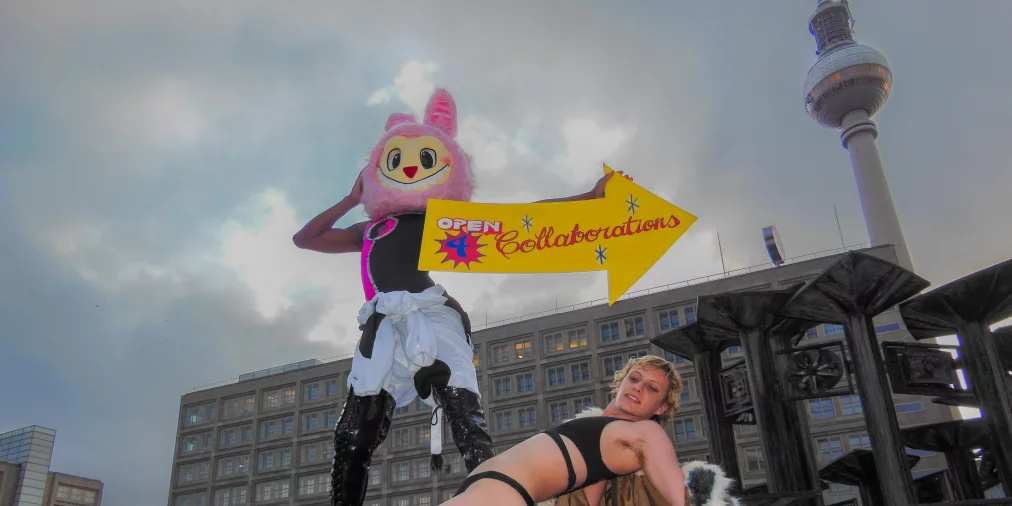 Eine Person in einem rosa Maskottchen-Kostüm steht erhöht am Brunnen, hält ein gelbes Pfeilschild mit „OPEN 4 Collaborations“. Davor posiert eine Person in schwarzer Unterwäsche und transparenten High Heels am Beckenrand. Im Hintergrund sind ein Gebäude und der Berliner Fernsehturm zu sehen.