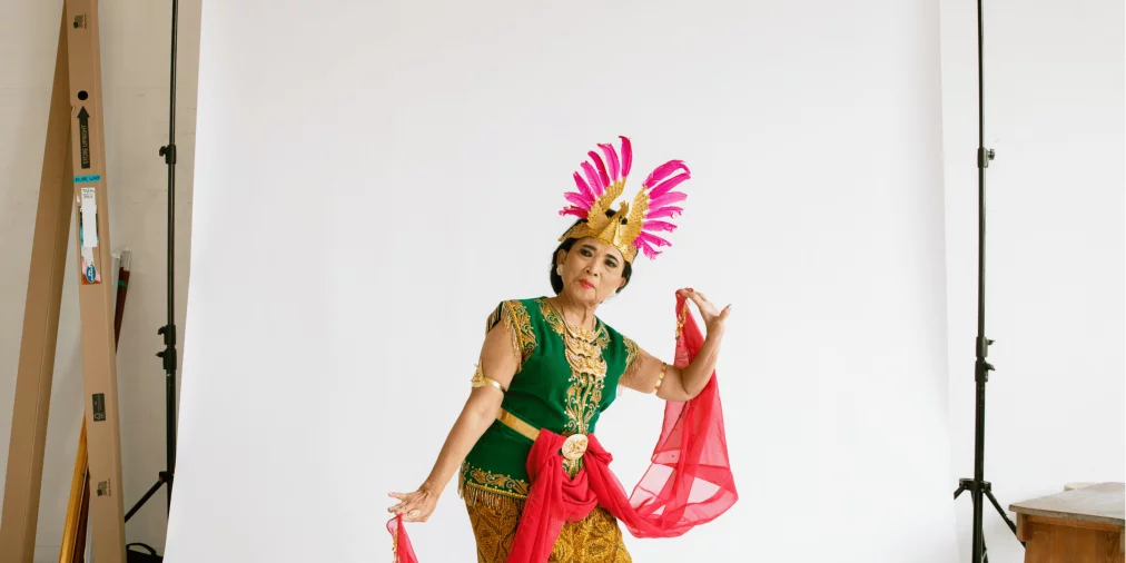 Eine ältere Tänzerin in farbenfrohem traditionellen Kostüm mit grünem Oberteil, goldenen Mustern und pinkem Kopfschmuck posiert vor einem weißen Studiohintergrund. Sie hält ein langes pinkes Tuch in beiden Händen und steht barfuß.