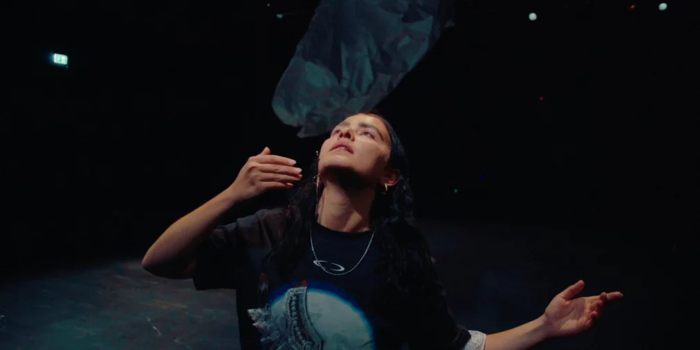 Eine Performer*in steht im Vordergrund und hebt den Blick suchend nach oben zum schwebenden, tuchartigen Objekt; ihre Arme sind leicht geöffnet in einer gestischen Bewegung.