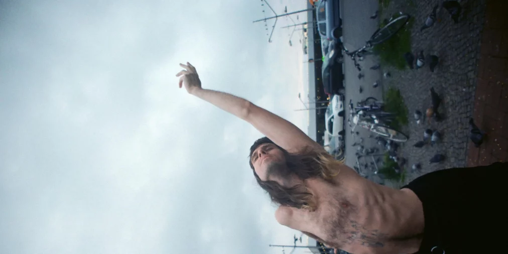 Eine oben unbekleidete Person mit langen Haaren steht zwischen Fahrrädern und Tauben und streckt einen Arm weit nach oben, den Kopf nach hinten gelehnt. Autos, Laternen und ein grauer Himmel sind im Hintergrund erkennbar.