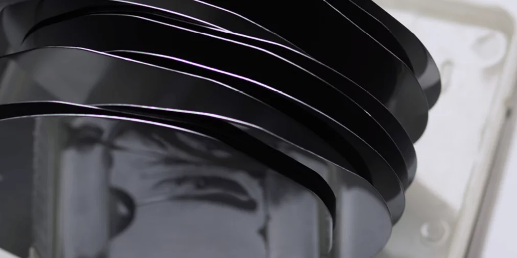 Workshop_3Mehrere übereinanderliegende schwarze Siliziumwafer in einer transparenten Halterung; ihre spiegelnden Oberflächen werfen weiche Reflexionen und verzerren Formen im Hintergrund.