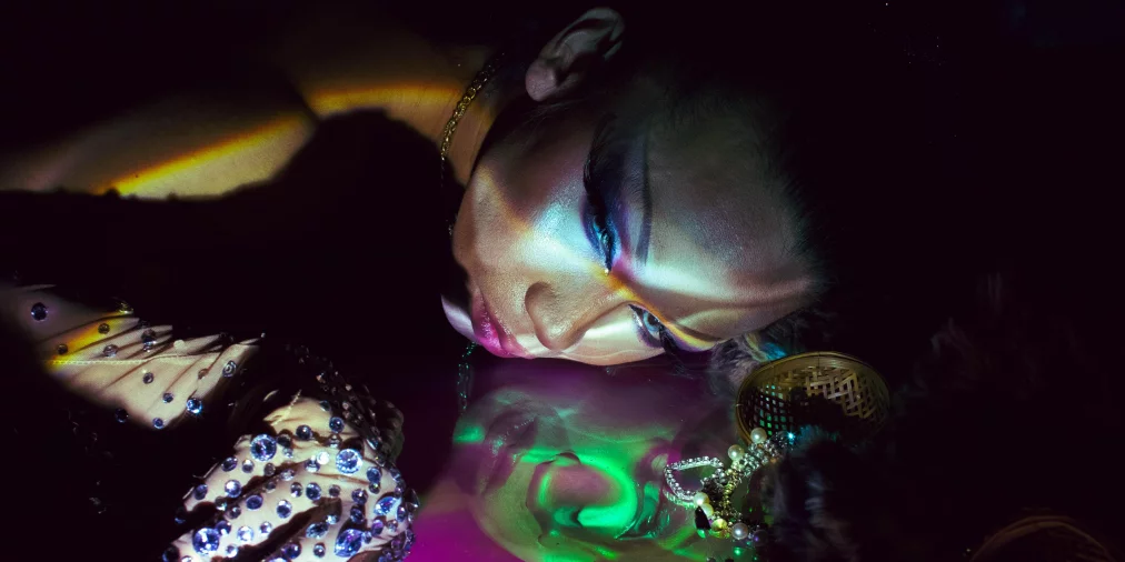 Eine Person liegt seitlich auf einer spiegelnden Fläche, das Gesicht von bunten Lichtprojektionen überzogen. Schmucksteine auf dem Handschuh funkeln, während sich das Gesicht klar in der glänzenden Oberfläche spiegelt.