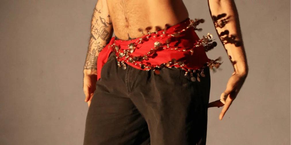 Nahaufnahme eines tanzenden Oberkörpers, bekleidet mit einer schwarzen Hose und einem roten Hüfttuch mit glänzenden Münzen und Perlen, die beim Bewegen schimmern. Tätowierte Arme rahmen die Szene, während warmes Licht Schatten auf den Körper wirft.