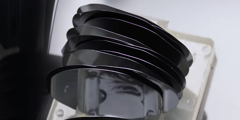 Several stacked black silicon wafers in a transparent holder; their reflective surfaces cast soft reflections and distort shapes in the background.