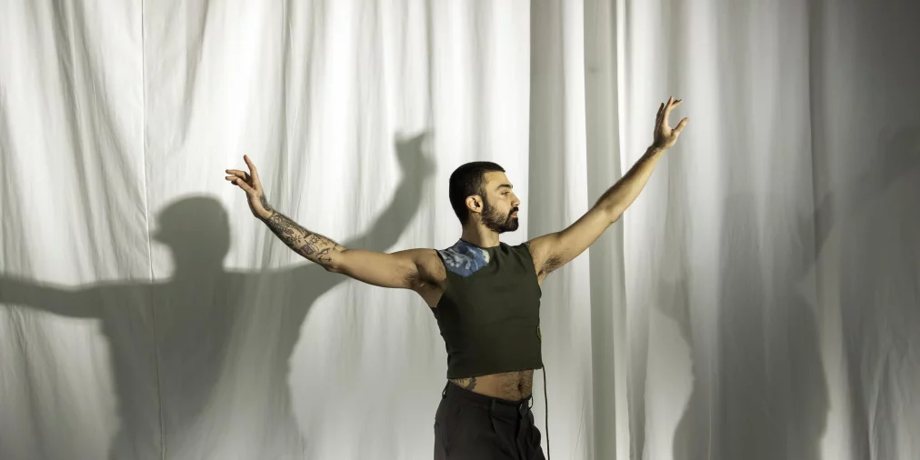 Eine Person steht seitlich vor einem weißen Vorhang und breitet die Arme weit aus, sodass ein großer Schatten hinter ihnen entsteht. Die Person trägt ein ärmelloses olivgrünes Oberteil und dunkle Hose. Der Körper ist in einer ruhigen, ausdrucksstarken Pose.