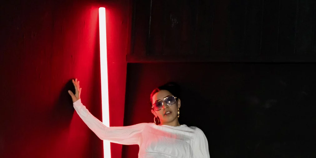 Eine Person in weißem Outfit steht breitbeinig in einem dunklen Raum. Eine vertikale rote Lichtleiste leuchtet hinter der Person, der linke Arm ist seitlich ausgestreckt, die rechte Hand liegt am Oberschenkel. Auf dem Boden liegen silberne, bandartige Streifen.