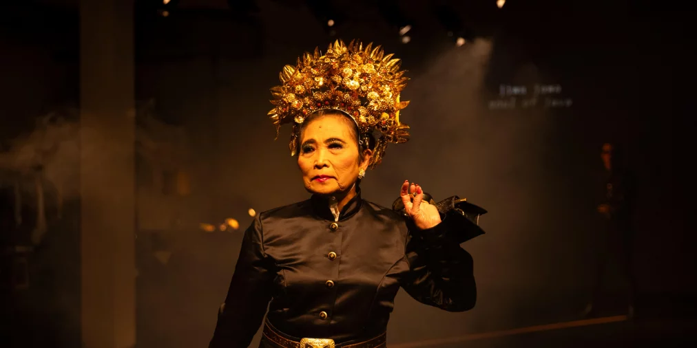 Eine Person steht frontal auf der Bühne und trägt ein schwarzes Kostüm mit goldenen Details sowie eine große, goldene Kopfbedeckung. Warmes Licht fällt von oben, der Hintergrund ist dunkel und leicht verraucht.