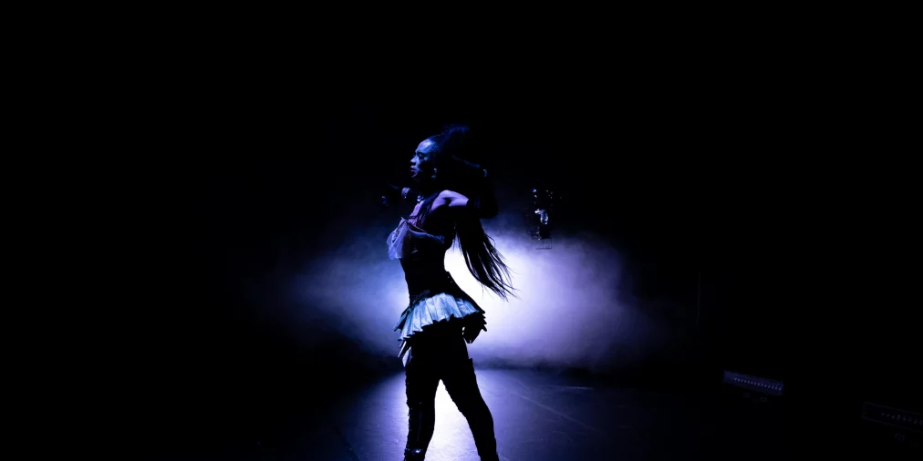 Eine Person steht seitlich auf einer dunklen Bühne, im Gegenlicht von blauem Licht und Nebel. Die Silhouette hebt sich klar vom Hintergrund ab, während lange Haare nach hinten fallen und hohe Plateauschuhe sichtbar sind.
