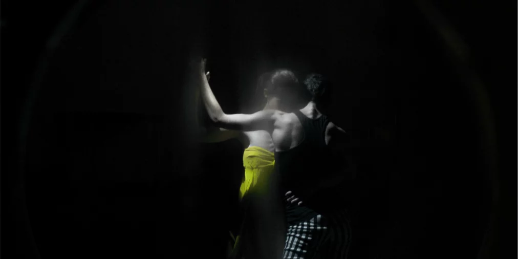 Zwei Personen stehen eng beieinander im Dunkeln, von hinten gesehen; eine trägt ein leuchtend gelbes Kleid, die andere ein schwarzes Oberteil und gestreifte Hose, beide teilweise von Licht und Schatten umhüllt.