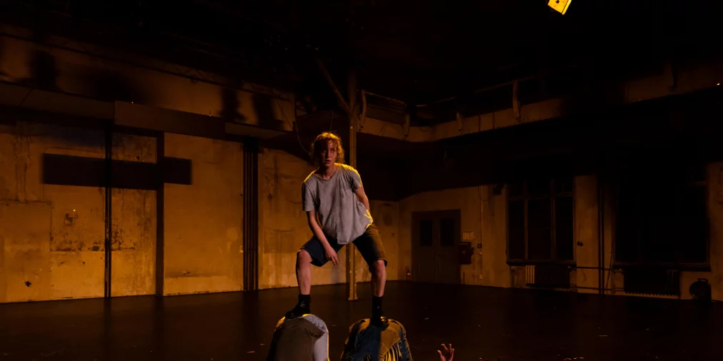 Eine Person steht mit gespreizten Beinen erhöht auf zwei anderen Personen, die sich nach unten abstützen. Der Raum wirkt industriell und leer, das Licht ist warm und konzentriert auf die Körper.