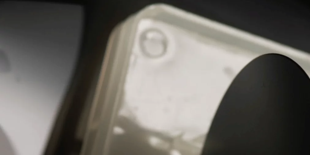 Unscharfe Nahaufnahme eines hellen Kunststoffgehäuses mit Kanten und Schrauben, teilweise verdeckt von einem dunklen, gebogenen Objekt im Vordergrund. Das Licht erzeugt weiche Spiegelungen und Schatten.