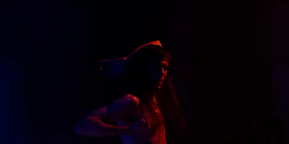 Eine obenrum nackte Person bewegt sich in starkem Rot- und Blaulicht. Ein Arm ist hinter den Kopf geführt, der andere angewinkelt vor dem Körper. Der Hintergrund ist dunkel, die Körperhaltung wirkt angespannt und konzentriert.