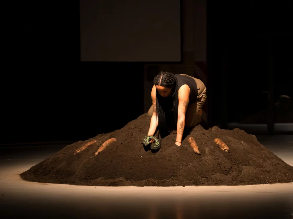 Eine Performer*in kniet auf einem großen Erdhügel und gräbt mit den Händen in der Erde. Das Licht fällt gezielt auf ihre Körperhaltung, während der restliche Raum dunkel bleibt. Hinter ihr sind die Umrisse einer Projektionsfläche erkennbar.
