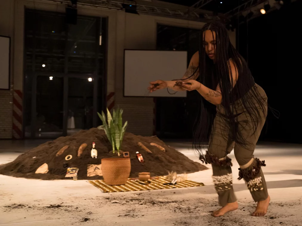 Eine Performer*in bewegt sich barfuß über den Boden und streut Erde aus ihren Händen. Hinter ihr liegt ein Erdhügel mit arrangierten Objekten, darunter eine Pflanze in einem Korb, Flaschen, Holzstücke und kleinere Artefakte. Die Szene ist atmosphärisch ausgeleuchtet.