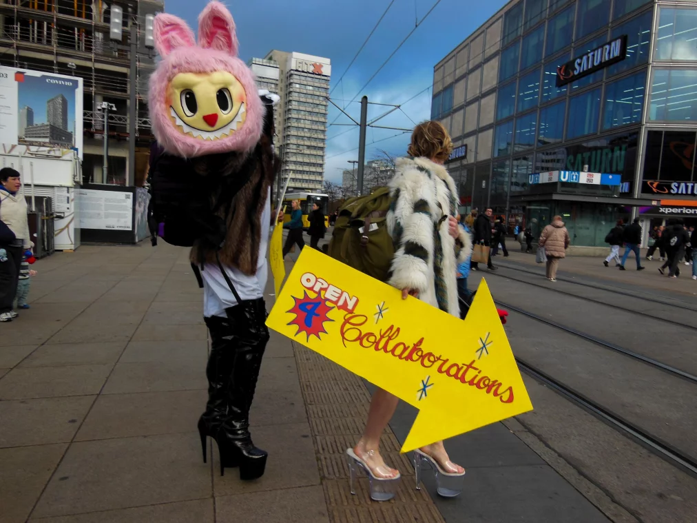 Zwei Personen im Stadtraum, eine trägt ein großes rosa Maskottchen-Kostüm mit Hasenohren und sehr hohe schwarze Stiefel. Die andere Person trägt einen langen, gemusterten Kunstpelzmantel und transparente Plateau-Heels und hält ein großes gelbes Pfeilschild mit der Aufschrift „OPEN 4 Collaborations“. Passant*innen bewegen sich im Hintergrund über den Platz.