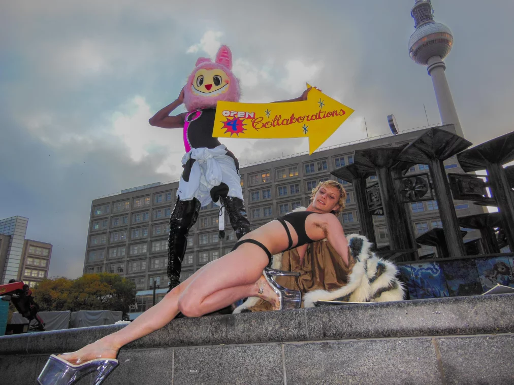 Eine Person in einem rosa Maskottchen-Kostüm steht erhöht am Brunnen, hält ein gelbes Pfeilschild mit „OPEN 4 Collaborations“. Davor posiert eine Person in schwarzer Unterwäsche und transparenten High Heels am Beckenrand. Im Hintergrund sind ein Gebäude und der Berliner Fernsehturm zu sehen.