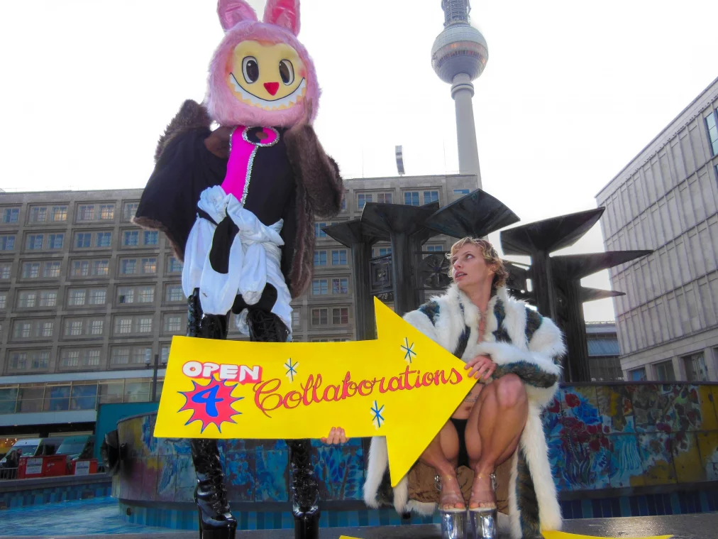 Eine Person im rosa Maskottchen-Kostüm steht vor dem Brunnen am Alexanderplatz und hält gemeinsam mit einer Person im gestreiften Kunstpelzmantel ein großes gelbes Pfeilschild, auf dem „OPEN 4 Collaborations“ steht. Beide tragen sehr hohe Plateau-Absätze. Hinter ihnen ragt der Berliner Fernsehturm in den Himmel.