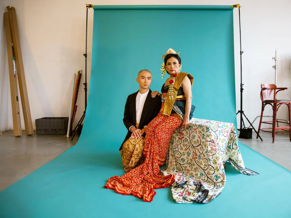 Eine ältere Tänzerin in opulentem golden-rotem Gewand sitzt auf einem mit gemustertem Stoff bedeckten Podest, während eine weitere Person neben ihr auf dem Boden sitzt. Beide tragen Elemente traditioneller Kleidung und blicken ruhig in Richtung Kamera.