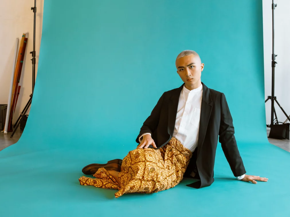 Eine Person sitzt lässig auf dem türkisfarbenen Studiohintergrund, trägt einen schwarzen Blazer, ein weißes Hemd und einen goldgemusterten Sarong. Der Blick ist direkt und ruhig in die Kamera gerichtet.