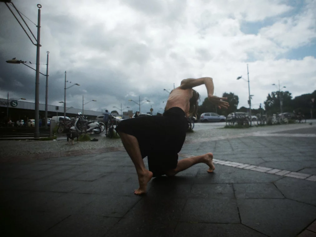 Eine oben unbekleidete Person bewegt sich in einer tiefen, dynamischen Tanzpose über einen nassen Platz. Fahrräder, Autos und ein wolkenverhangener Himmel bilden den Hintergrund.