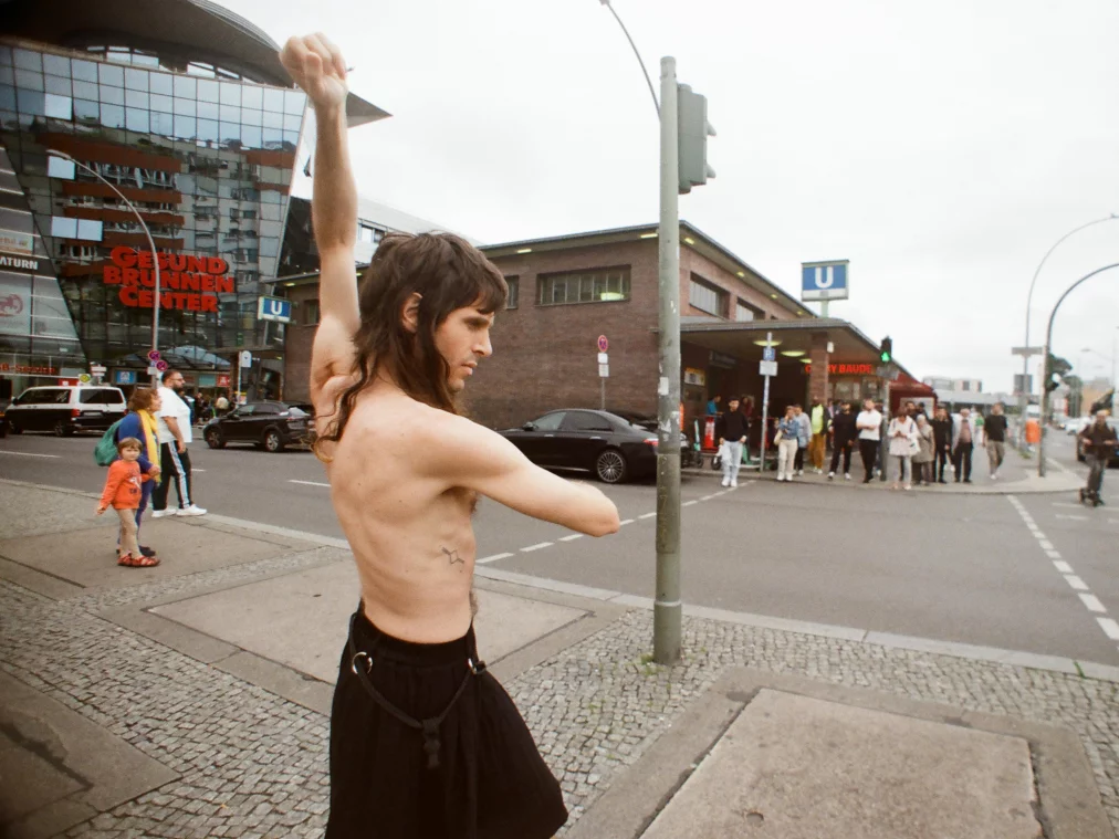 Eine oben unbekleidete Person mit langen Haaren steht auf einem Gehweg an einer Berliner Kreuzung und hebt einen Arm in einer tänzerischen Geste. Im Hintergrund sind das Gesundbrunnen-Center, eine U-Bahn-Station und vorbeigehende Menschen zu sehen.
