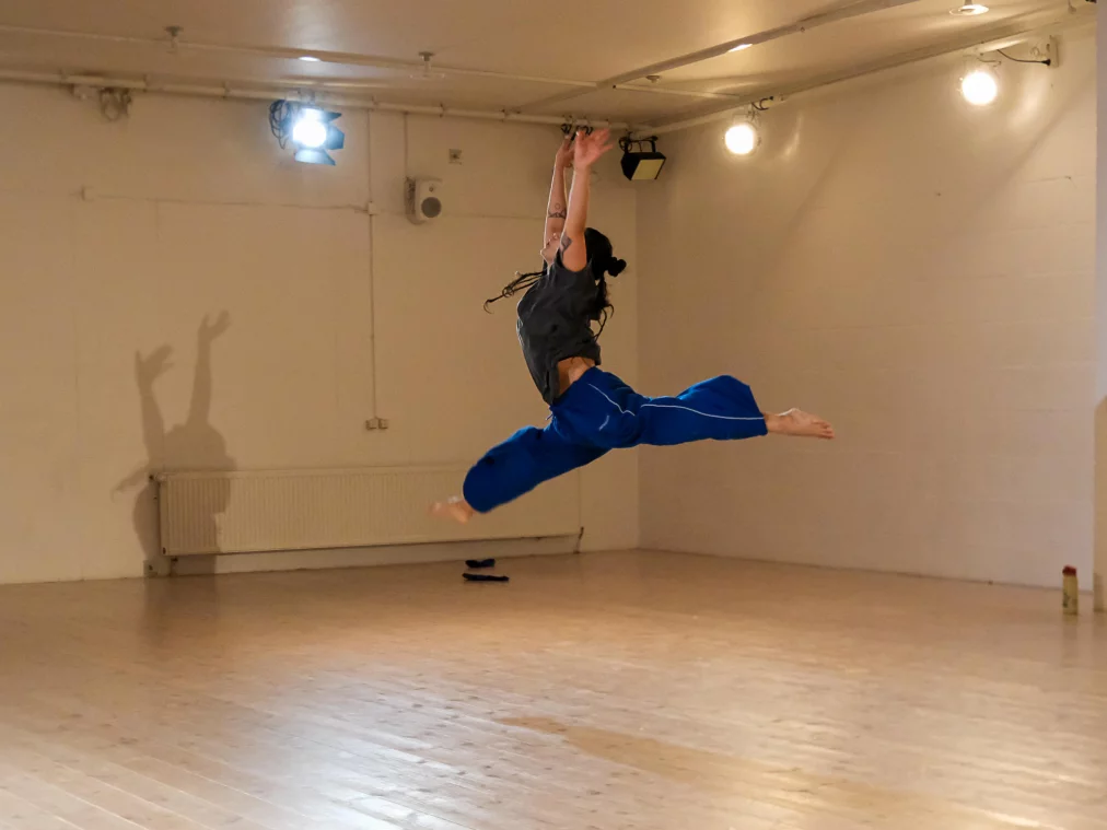 Eine tanzende Person springt weit durch den Raum, beide Beine in einem großen Spagat in der Luft. Die Arme sind nach oben gestreckt, das lange Haar bewegt sich im Sprung. Der helle Proberaum ist leer bis auf wenige Gegenstände am Rand.