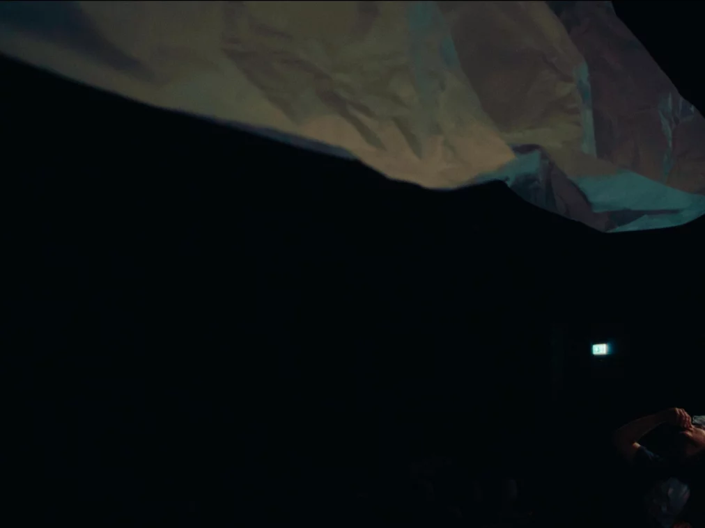 Eine Performer*in erhebt den Arm in einer fließenden Bewegung, während ein großes, schwebendes Tuch knapp über ihr im Scheinwerferlicht schimmert.