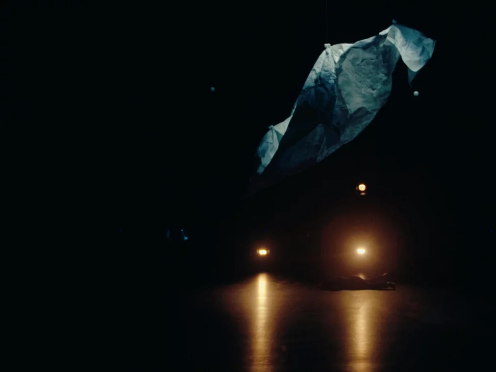 Ein großes, zerknittertes Tuch schwebt zentral über der relativ dunklen Bühne, darunter liegt eine Performer*in reglos im warmen Schein zweier Scheinwerfer.