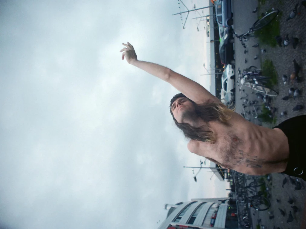 Eine oben unbekleidete Person mit langen Haaren steht zwischen Fahrrädern und Tauben und streckt einen Arm weit nach oben, den Kopf nach hinten gelehnt. Autos, Laternen und ein grauer Himmel sind im Hintergrund erkennbar.