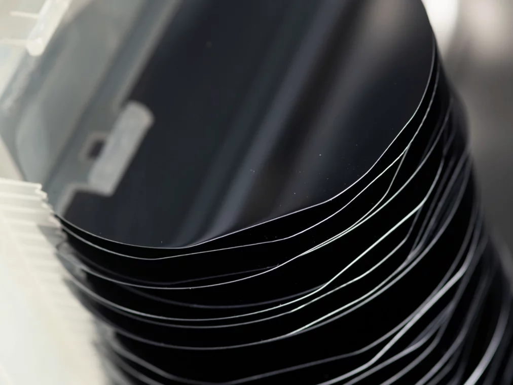 Detailaufnahme eines gestapelten Sets hochglänzender schwarzer Siliziumwafer in einer Kunststoffhalterung; die feinen, gebogenen Kanten der dünnen Scheiben bilden eine rhythmische Linienstruktur.