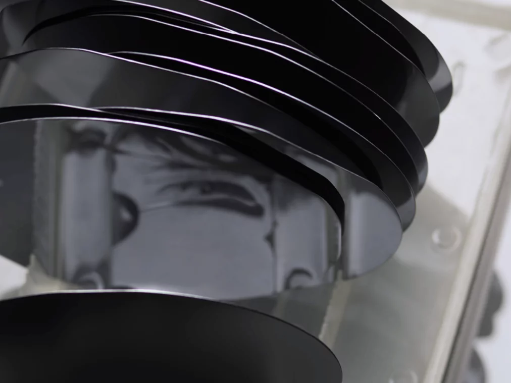 Workshop_3Mehrere übereinanderliegende schwarze Siliziumwafer in einer transparenten Halterung; ihre spiegelnden Oberflächen werfen weiche Reflexionen und verzerren Formen im Hintergrund.