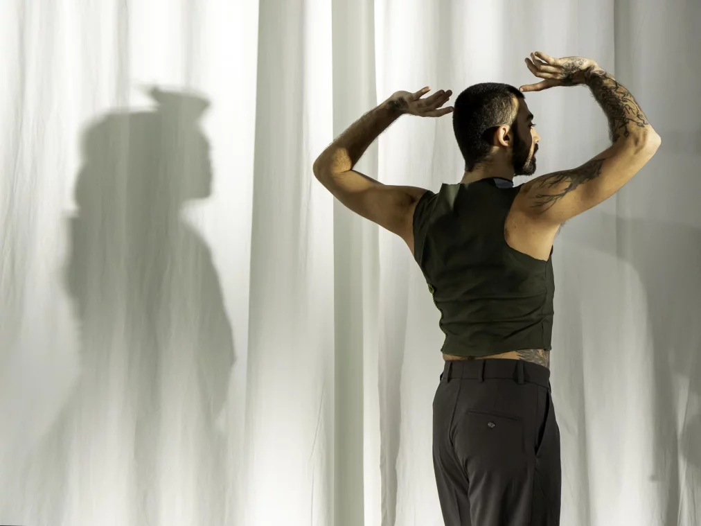 Eine Person steht mit dem Rücken zur Kamera vor weißen Vorhängen. Die Arme sind erhoben und angewinkelt, sodass ein deutlicher Schatten der Pose auf dem Vorhang erscheint. Die Person trägt ein ärmelloses dunkelgrünes Oberteil und dunkle Hose und hat sichtbare Armtätowierungen.