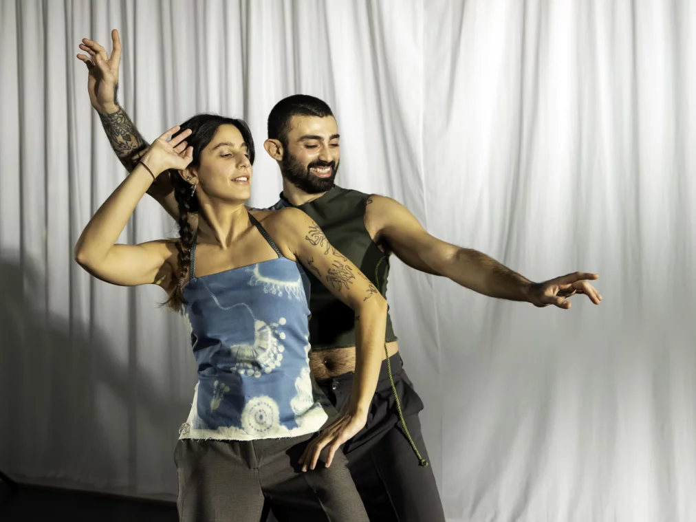 Zwei Personen stehen nah beieinander vor weißen Vorhängen und bewegen sich tanzend. Beide haben die Arme erhoben; eine Person zeigt nach vorne, die andere hat eine Hand am Kopf. Sie tragen ärmellose Oberteile und dunkle Hosen und wirken in einen gemeinsamen Bewegungsfluss vertieft.