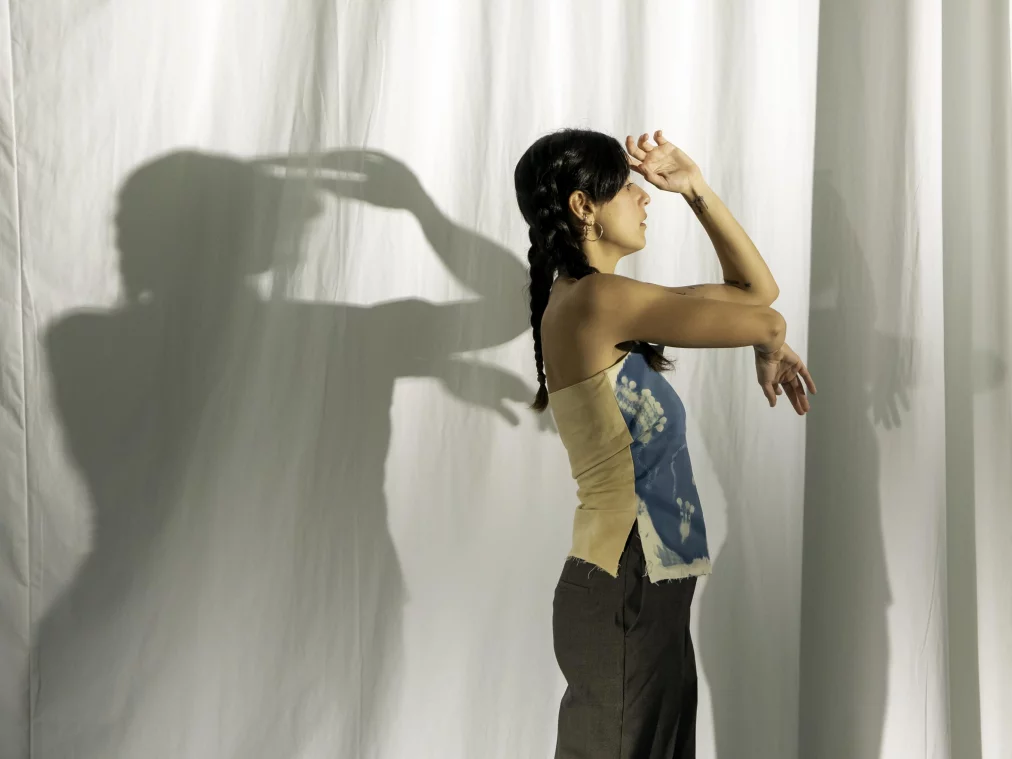 Eine Person im Profil steht vor einem weißen Vorhang und hebt eine Hand schützend über die Augen, während die andere leicht angewinkelt ist. Der Schatten der Person fällt deutlich auf den Vorhang. Sie trägt ein zweifarbiges, ärmelloses Oberteil und dunkle Hose; das Haar ist zu zwei Zöpfen geflochten.