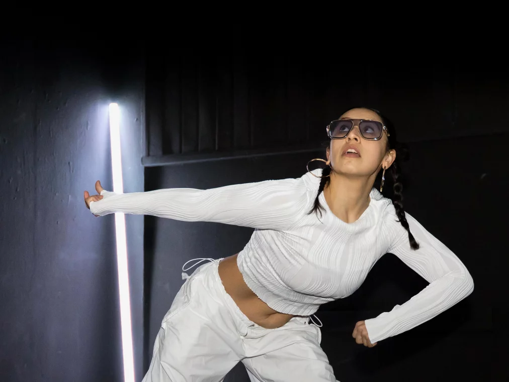 Eine Person in weißer Kleidung beugt den Oberkörper nach vorne und streckt beide Arme seitlich aus. Eine helle vertikale Lichtleiste befindet sich links im Bild, silberne Streifen verlaufen über den dunklen Boden.