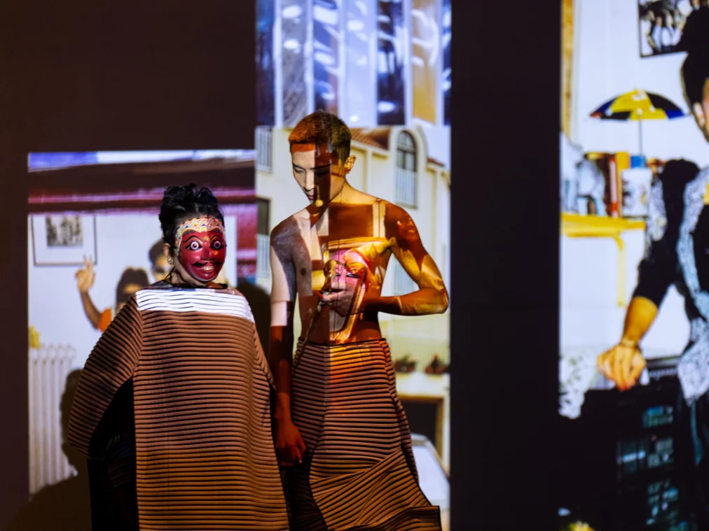 Zwei Personen stehen vor projizierten Bildern an einer Wand. Eine Person trägt eine Maske mit rotem Gesicht, die andere hält eine weitere Maske in den Händen. Die Projektionen zeigen Innenräume und Porträts.