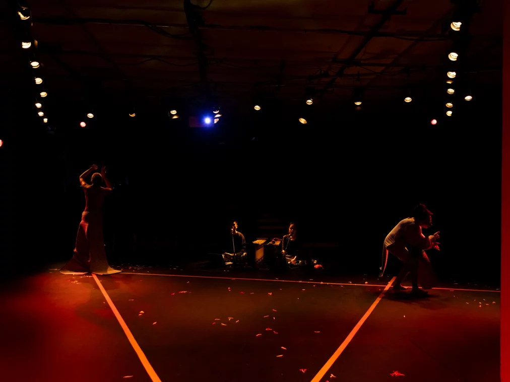 Weite Bühnenansicht mit mehreren Personen in unterschiedlichen Positionen. Zwei Personen bewegen sich an den Seiten, während drei Personen sitzend im Hintergrund zu sehen sind. Der Bühnenraum ist dunkel, mit geometrischen Lichtlinien auf dem Boden.