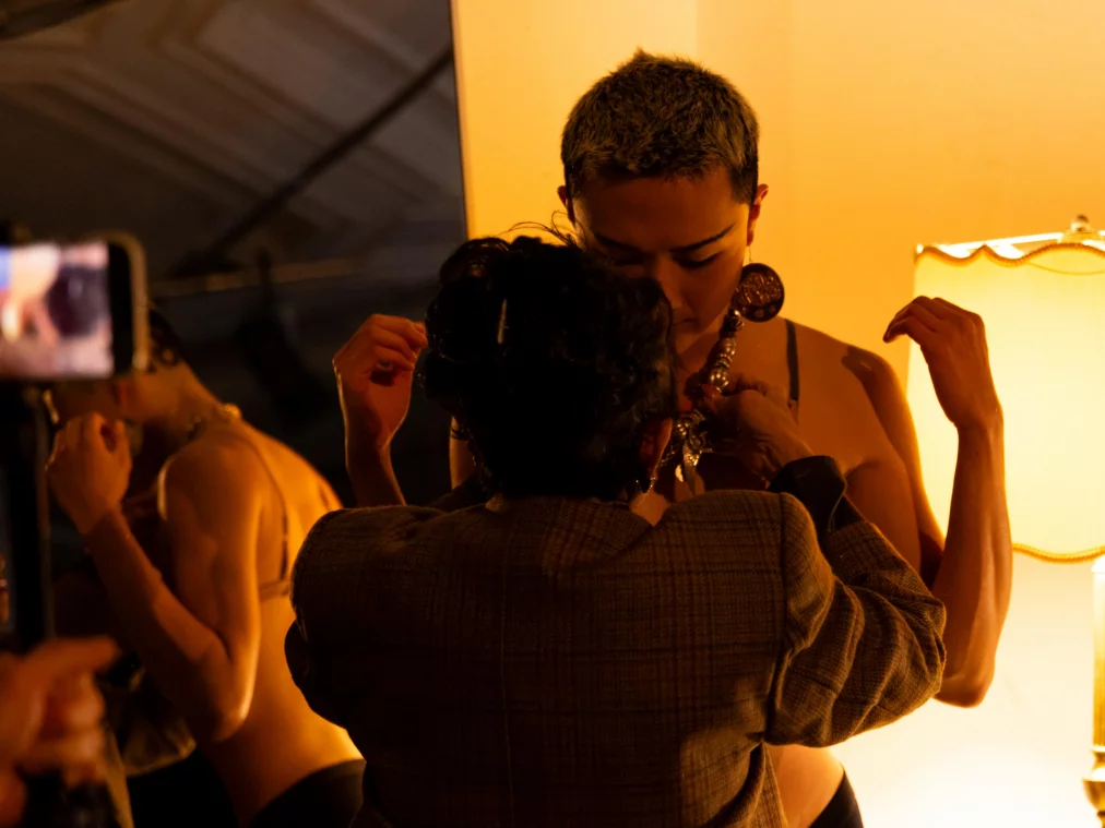 Zwei Personen stehen sich nah gegenüber in einem warm beleuchteten Innenraum. Eine Person richtet Schmuck am Hals der anderen Person. Ein Spiegel und eine Stehlampe sind im Hintergrund sichtbar.