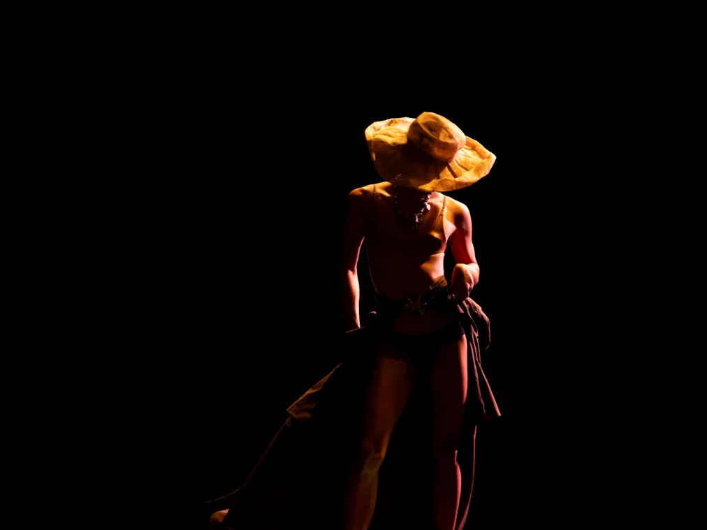 Eine einzelne Person steht nahezu nackt auf einer dunklen Bühne und trägt einen breiten Hut, der das Gesicht verdeckt. Der Körper ist seitlich beleuchtet, während der Hintergrund vollständig schwarz bleibt.