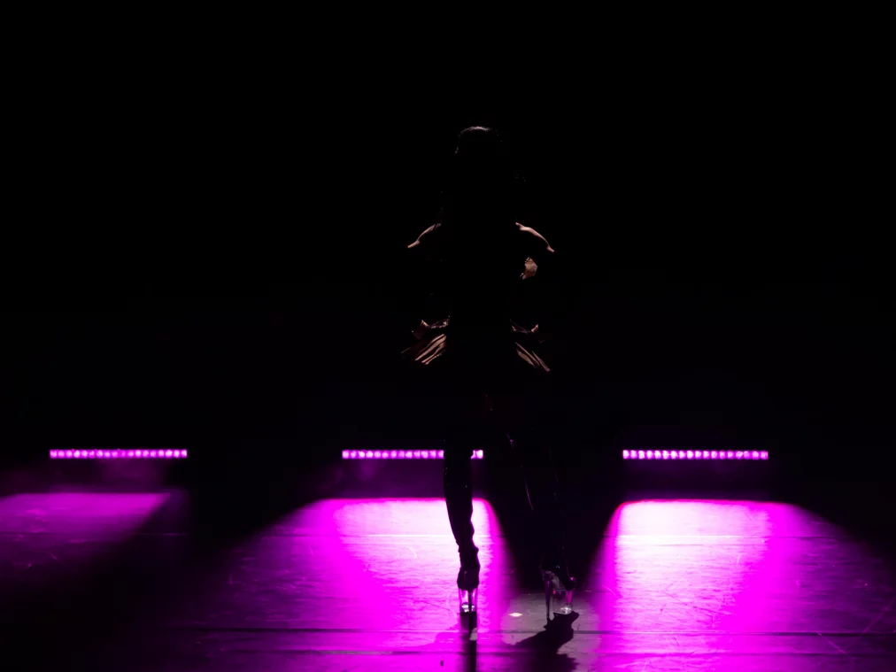 Die Silhouette einer Person steht frontal auf der Bühne, die Hände in die Hüften gestützt. Violettes Licht am Boden zeichnet die Konturen der Beine und der hohen Schuhe nach.