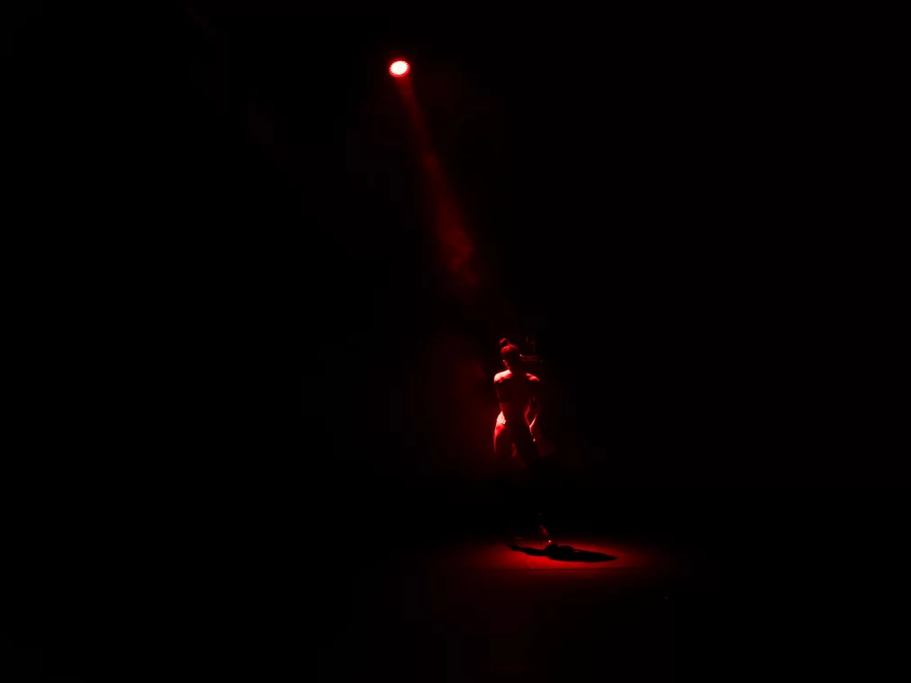 Eine Person steht allein im Zentrum einer dunklen Bühne unter einem einzelnen roten Scheinwerfer. Das rote Licht erzeugt eine dramatische Atmosphäre und hebt die aufrechte Haltung der Person hervor.