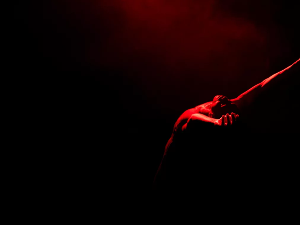 Eine Person beugt den Oberkörper nach hinten und streckt einen Arm weit nach oben. Rotes Licht fällt schräg von oben und hebt die gespannte Körperlinie vor dem schwarzen Hintergrund hervor.