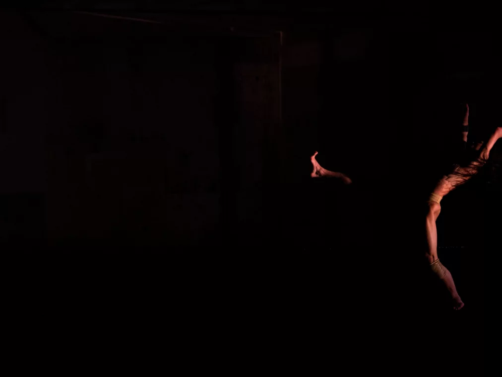 Eine Person bewegt sich seitlich durch den dunklen Bühnenraum, ein Arm und ein Bein weit ausgestreckt. Das Licht ist minimal und lässt den Körper nur teilweise aus der Dunkelheit hervortreten.