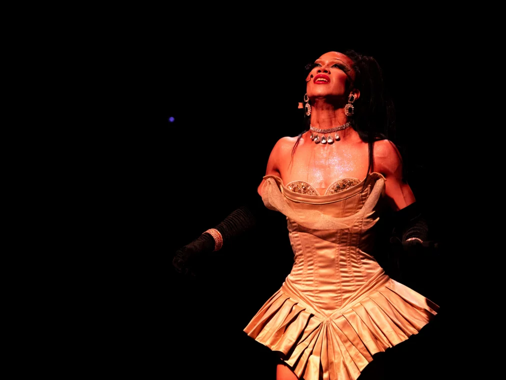 Eine Person im hellen Korsettkleid blickt nach oben, der Oberkörper glänzt im warmen Bühnenlicht. Schmuck und lange Handschuhe sind sichtbar, während der Hintergrund vollständig schwarz bleibt.