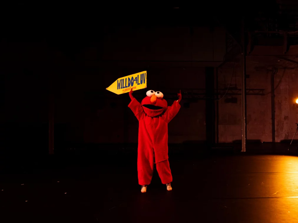 Eine Person im roten Ganzkörperkostüm steht mittig auf der Bühne und hält ein gelbes Schild über dem Kopf.