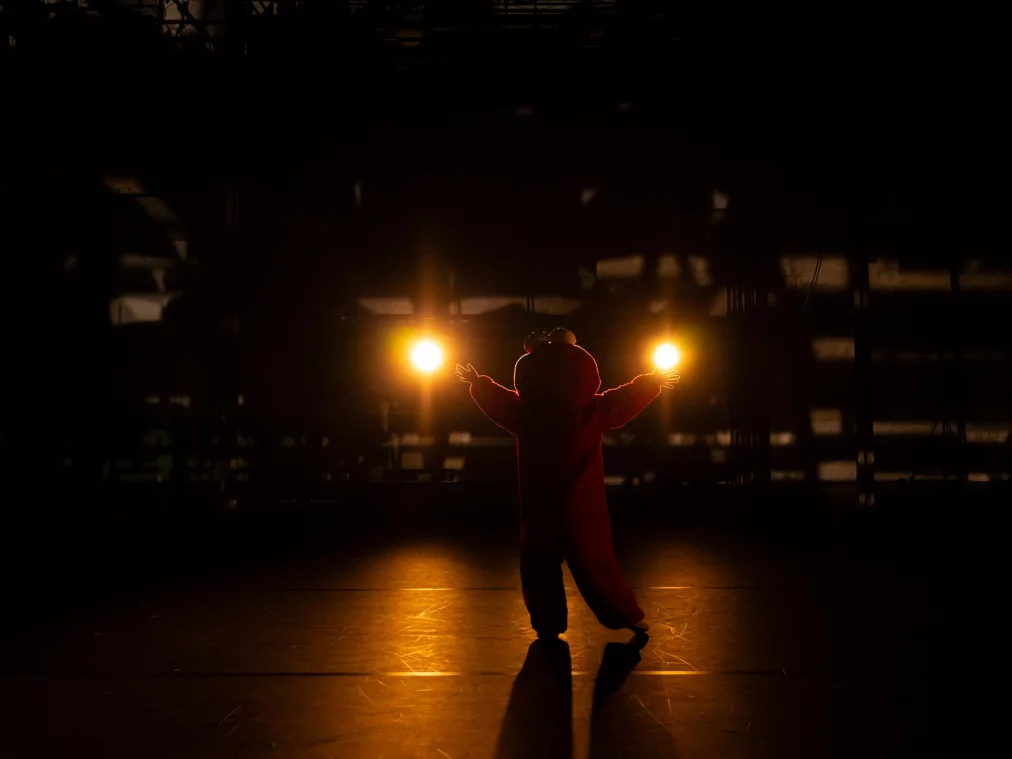 Eine Person im roten Ganzkörperkostüm steht mit ausgebreiteten Armen vor zwei hellen Scheinwerfern, als Silhouette im Gegenlicht.
