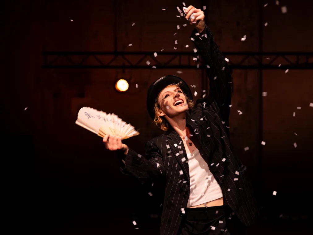 Eine Person im Anzug und Hut wirft Papier in die Luft und lächelt, warm beleuchtet vor dunklem Hintergrund.