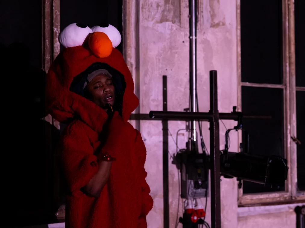 Nahaufnahme einer Person im roten Kostüm mit geöffnetem Mundbereich, das Gesicht wirkt erschöpft, im Halbdunkel.