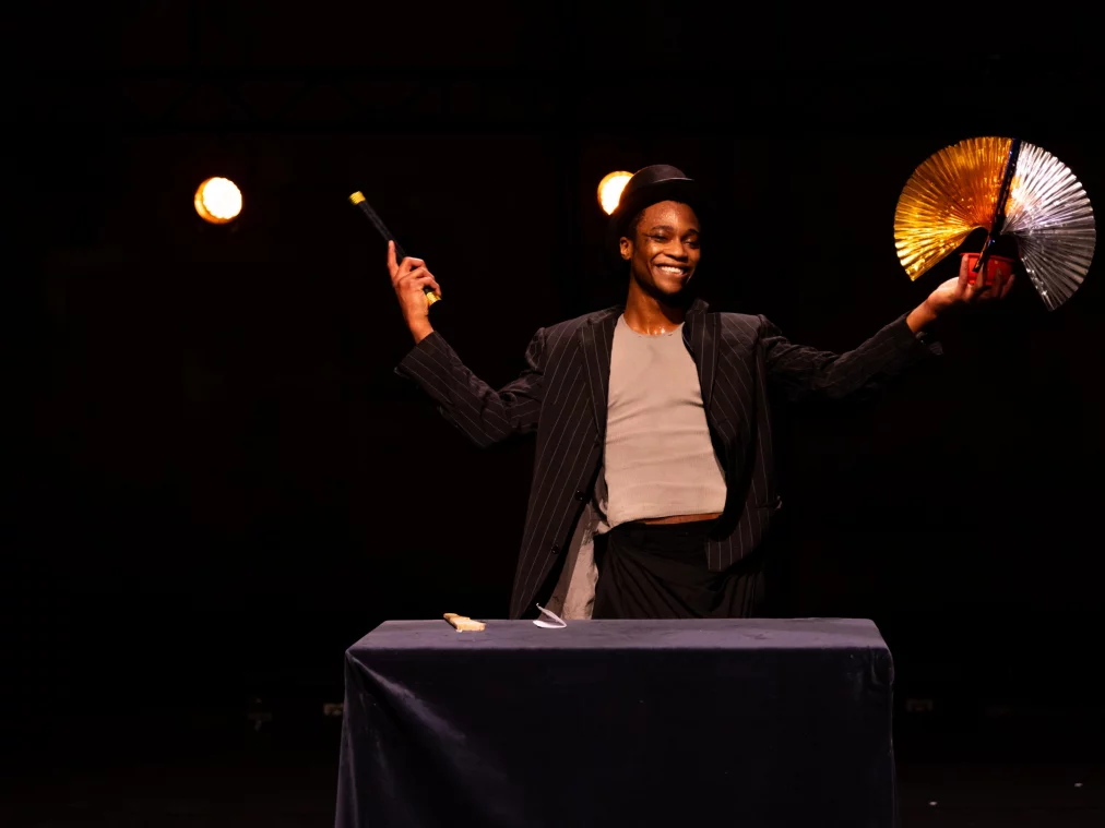 Eine Person im Anzug hält einen Fächer und einen Stab, lächelt breit und präsentiert sich frontal auf der Bühne.