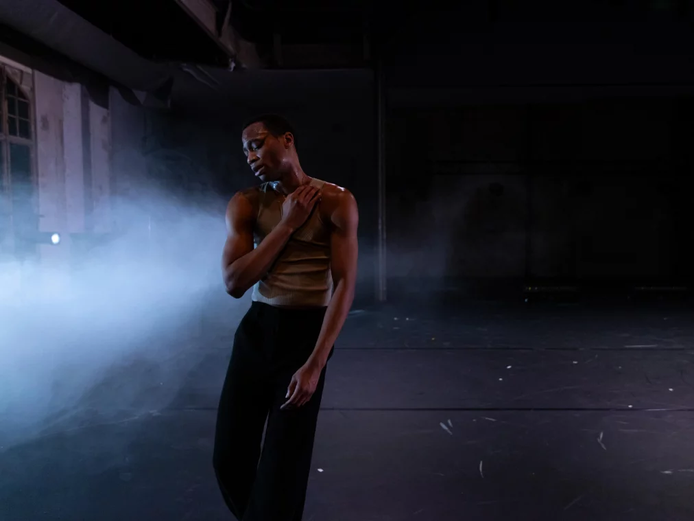 Eine Person steht im Bühnenraum und berührt die eigene Schulter, umgeben von Nebel und kühlem Licht.